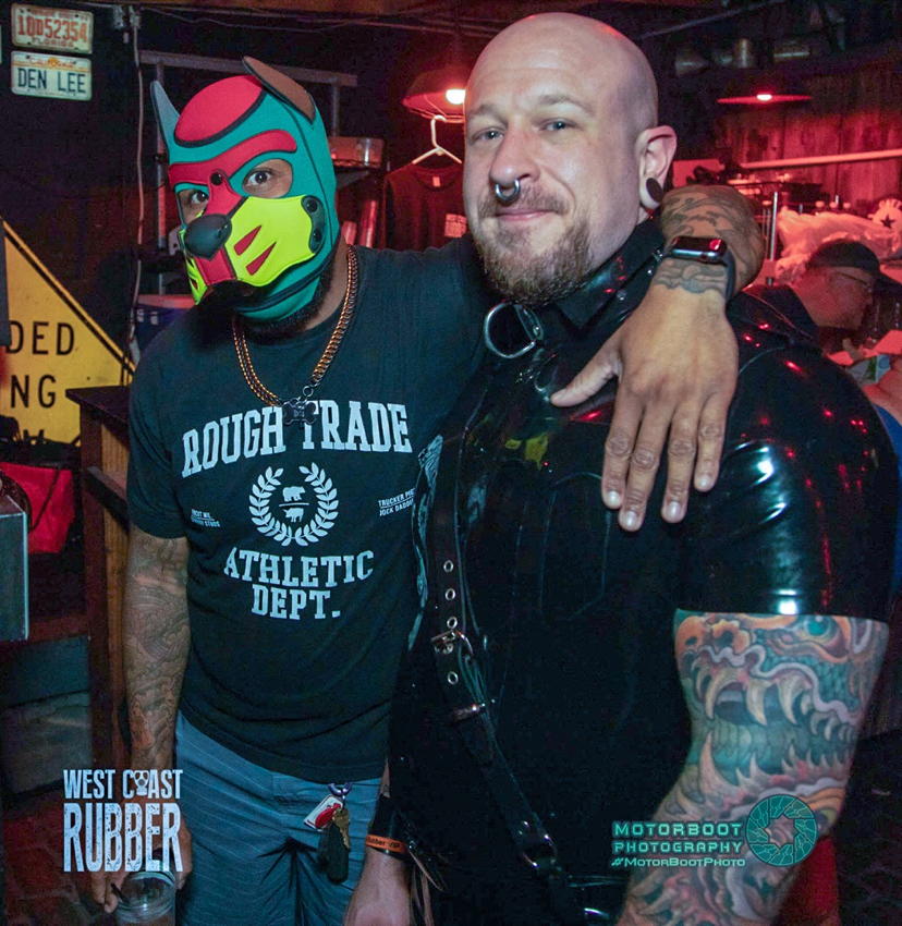 Black dude in garish pup hood has arm around stocky guy with nose ring and rubber shirting