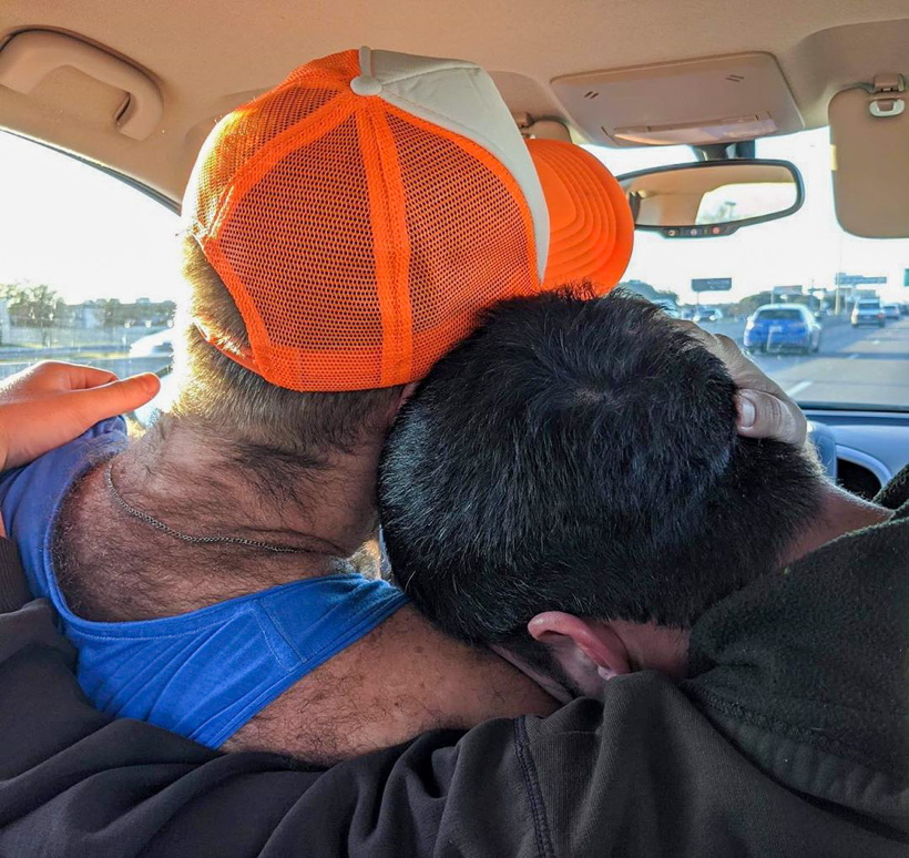 In a car, man in passengerseat buries his head in shoulder of driver, who wears an orange ballcap and a tank top revealing endless back and neck hair